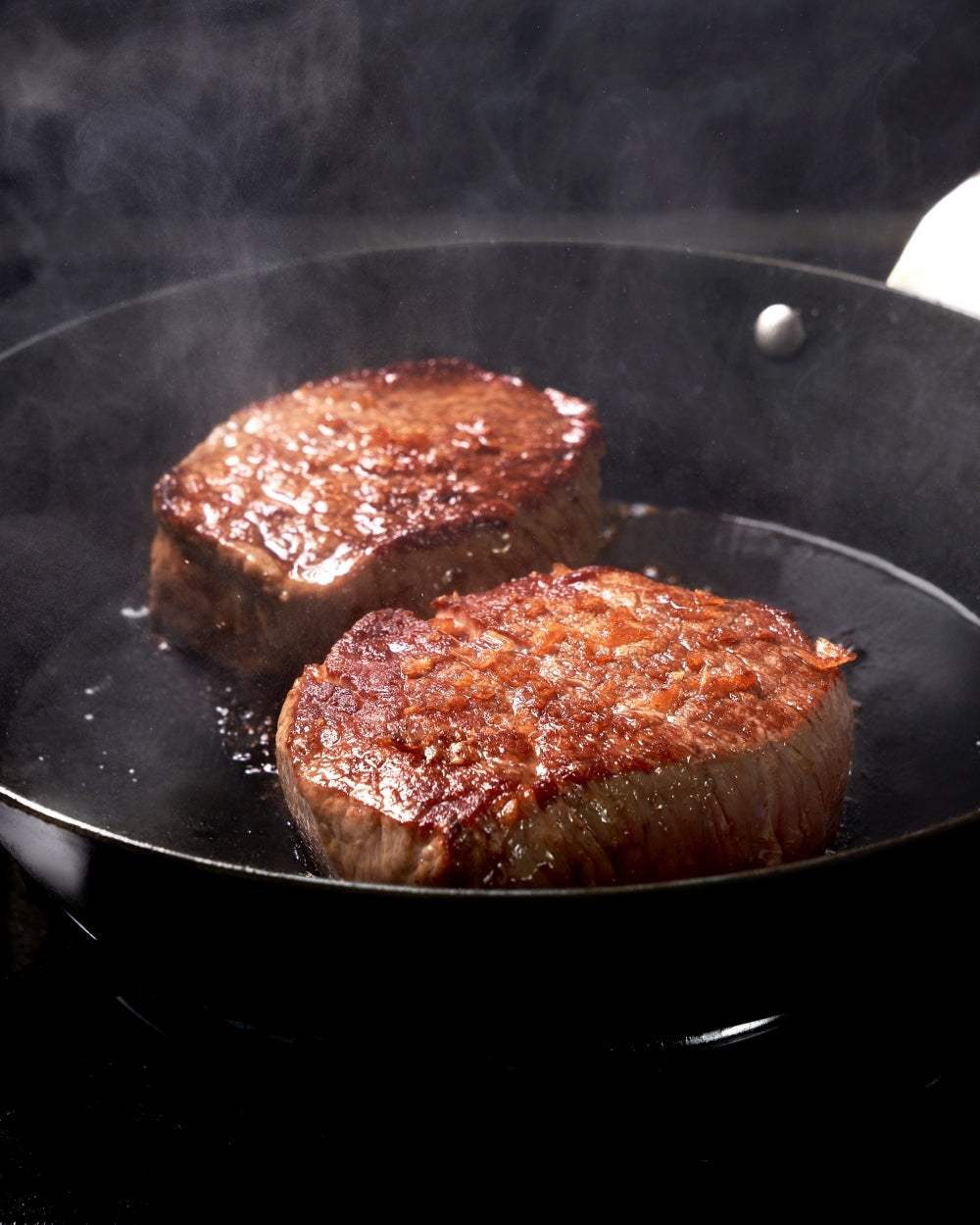 Two seared steaks cooking in a nonstick skillet with steam rising