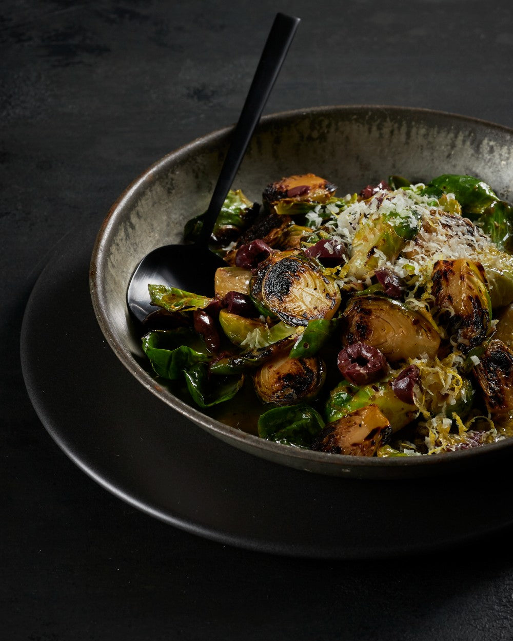 Charred Brussels sprouts with shaved cheese and olives in a rustic bowl on dark background