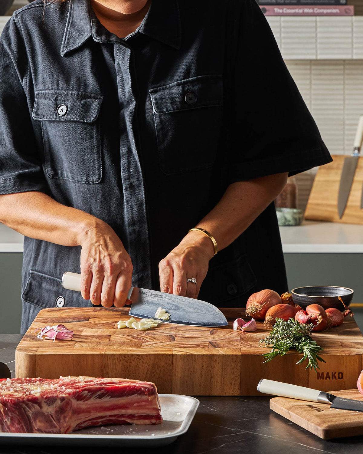 Person slicing garlic with a MAKO kitchen knife on a wooden cutting board, herbs and shallots nearby.