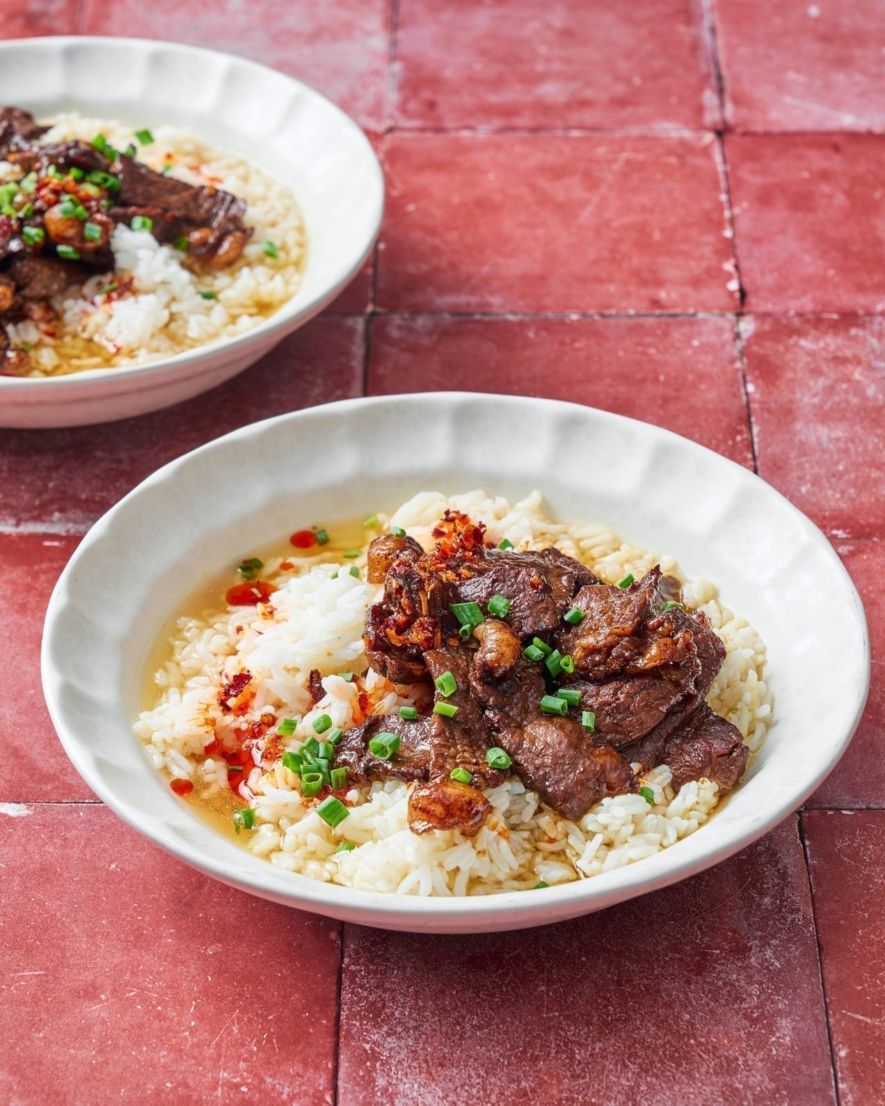 Brothy rice with caramelised steak, garlic ginger broth, spring onions and chilli oil in a white bowl on a red tiled surface