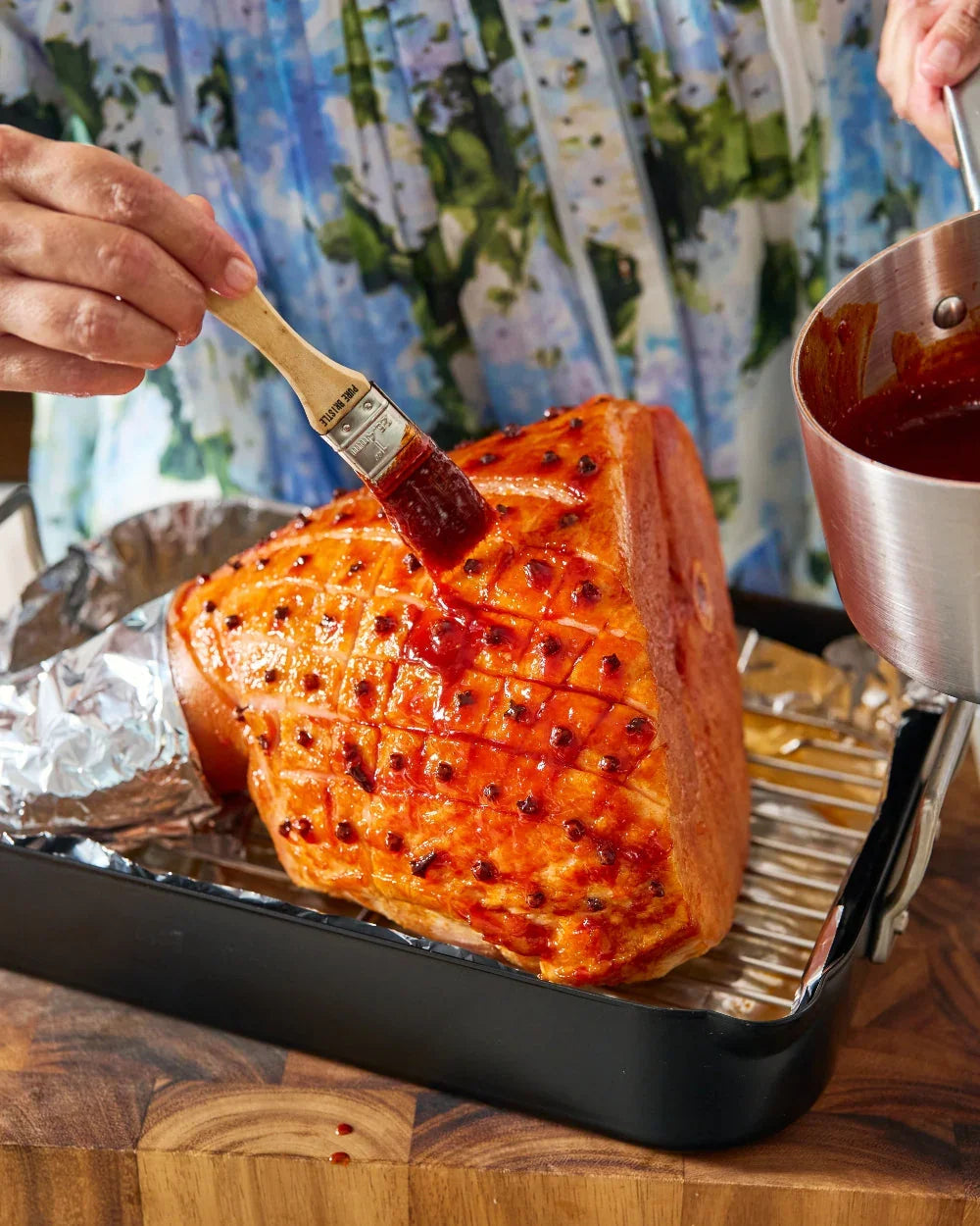 Glazed Christmas ham being brushed with sauce and studded with cloves in roasting pan.