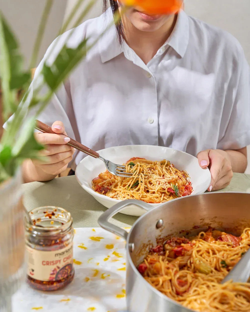 Woman eating spaghetti with tomato sauce and herbs, crispy chili jar and pot on table