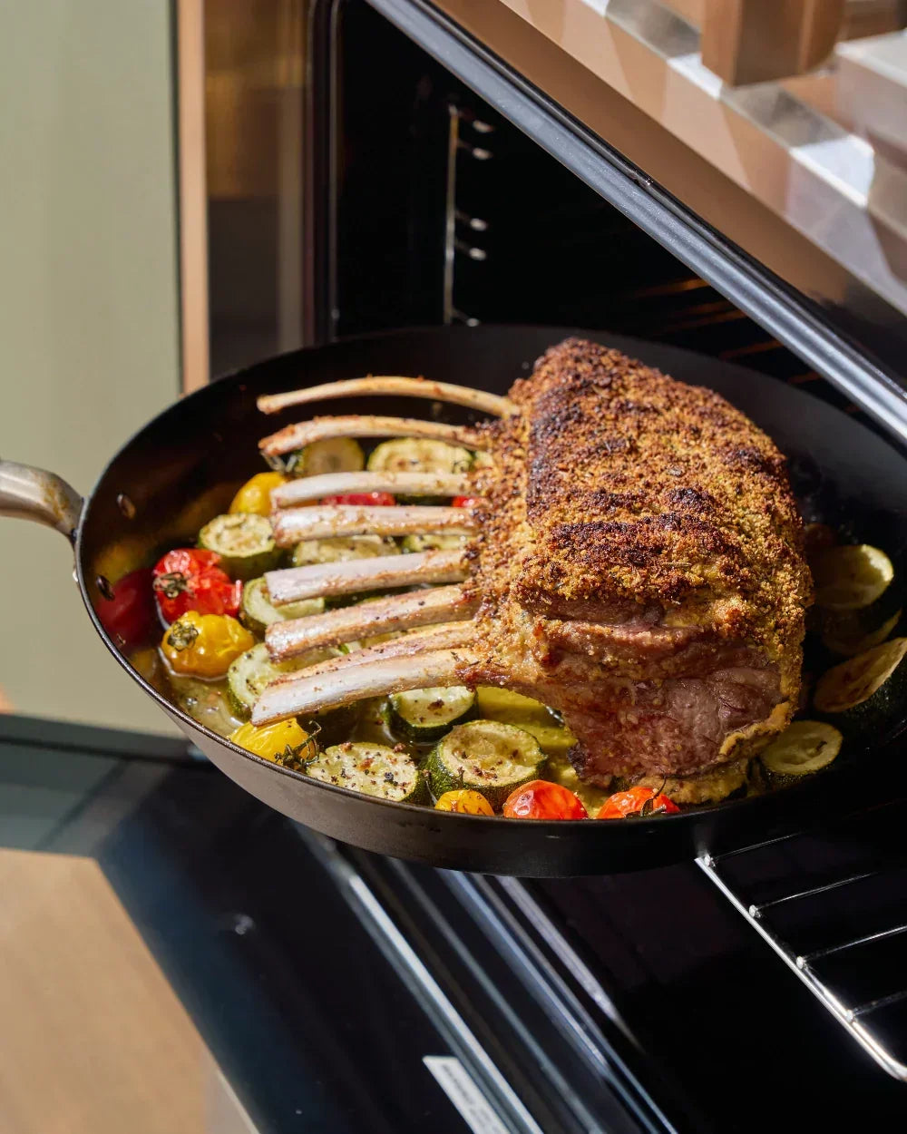 Herb-crusted rack of lamb roasting with zucchini and cherry tomatoes in a pan in the oven