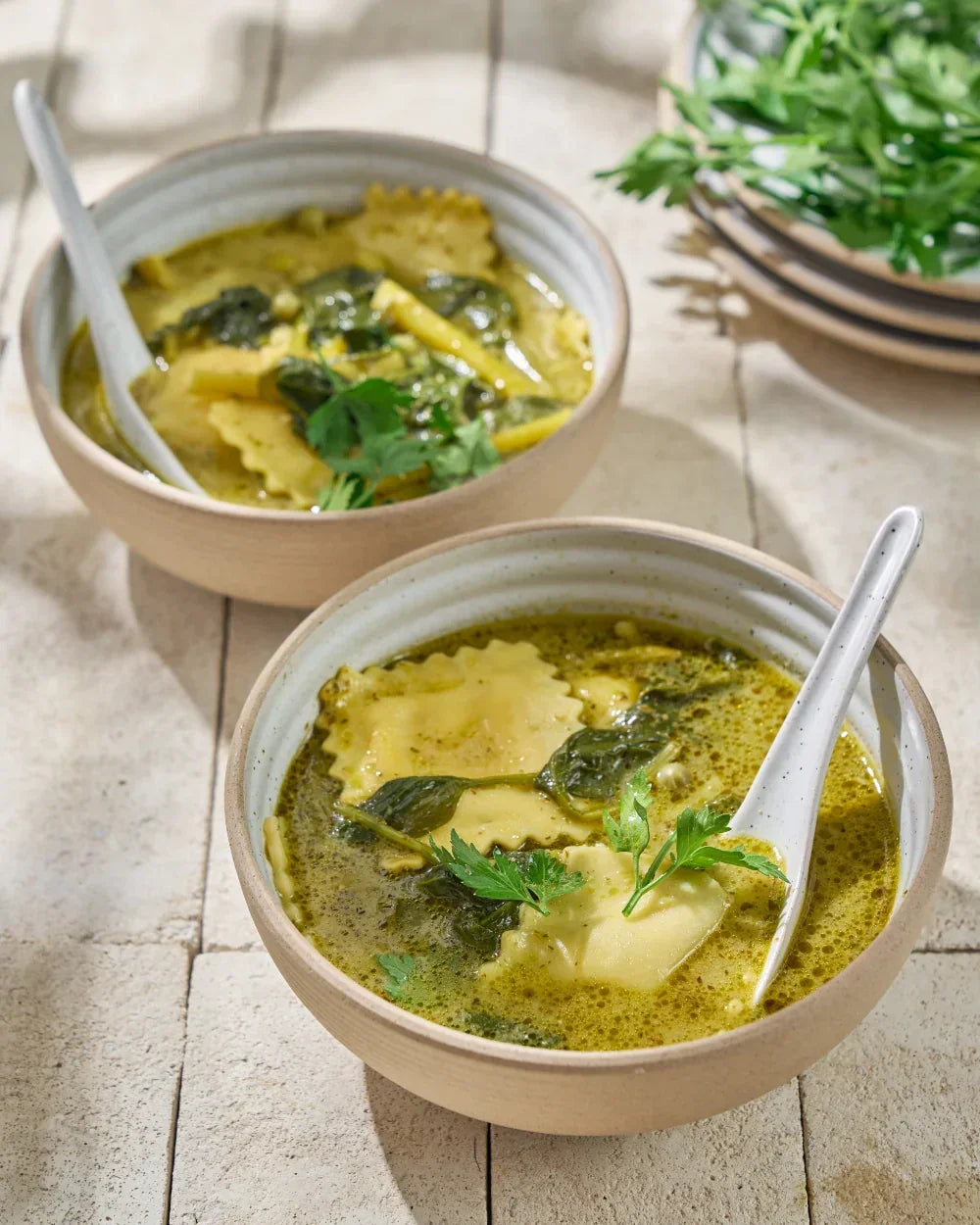 Two bowls of ravioli soup with herbs and greens, served in ceramic bowls on a sunny table.