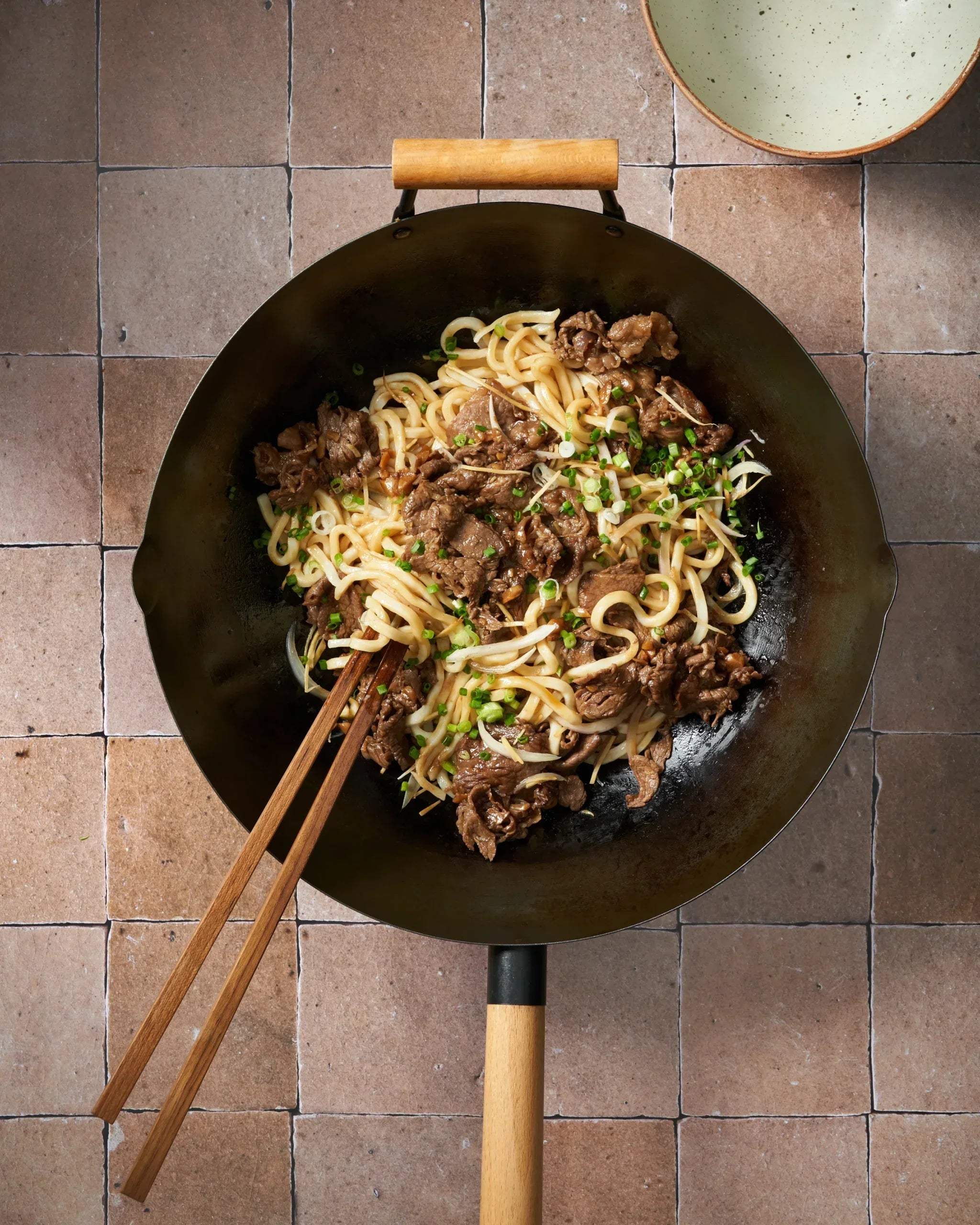 Mongolian beef udon noodles with green onions in a wok with wooden chopsticks on tiled surface