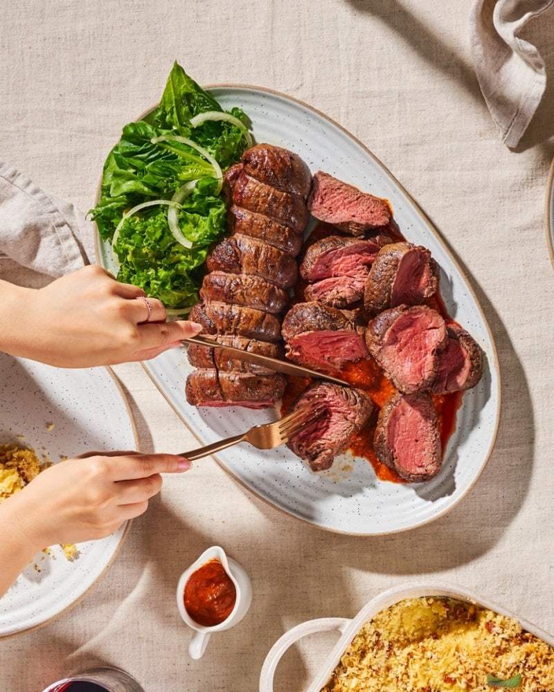 Sliced roast beef with salad on a white platter, hands carving, table setting with sauce