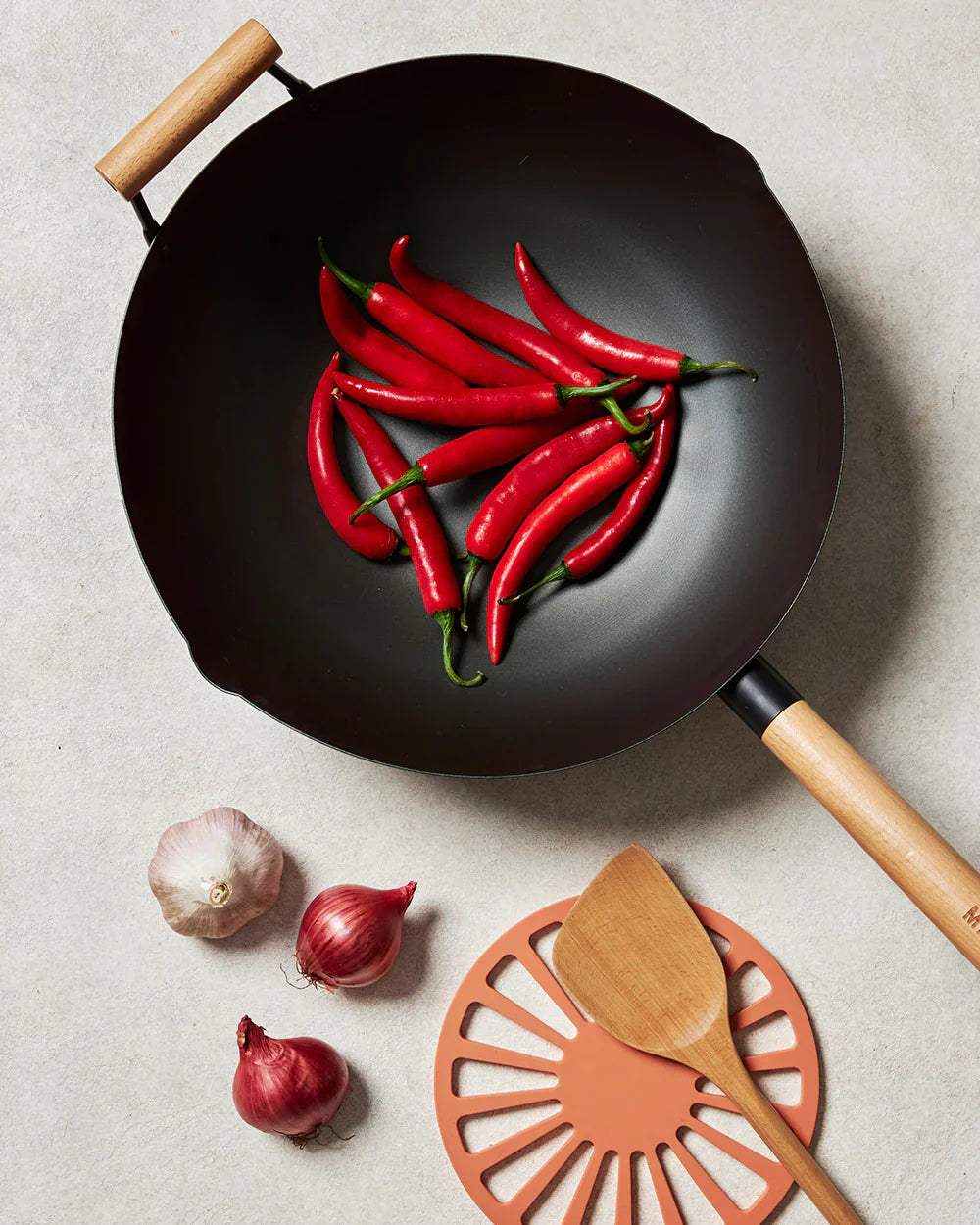 Black nonstick wok with red chili peppers, garlic, shallots, wooden spatula, and trivet on countertop