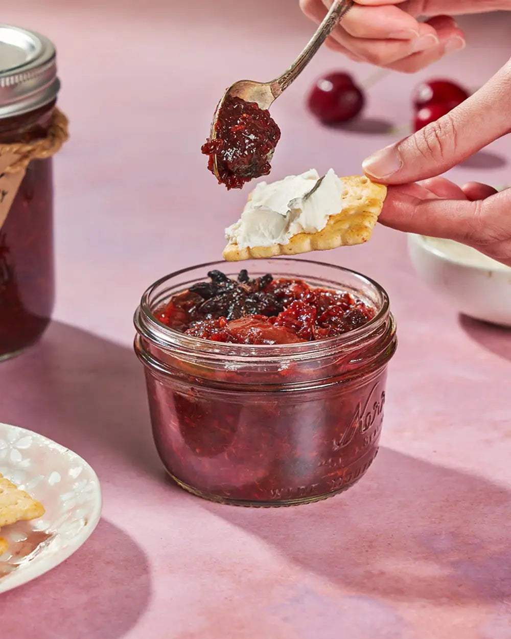 Cherry jam in a glass jar with a hand spreading it on a cracker with cream cheese