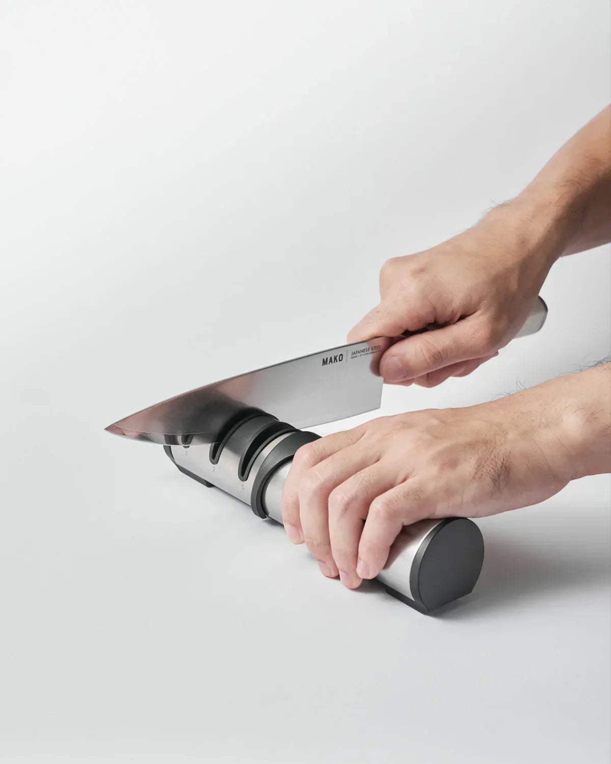 Person sharpening a Mako Japanese steel kitchen knife using a three-stage knife sharpener on a white surface