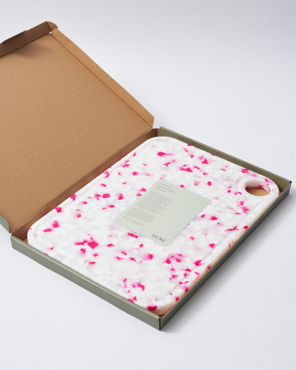 Speckled pink and white cutting board with handle in an open box, minimalist style.