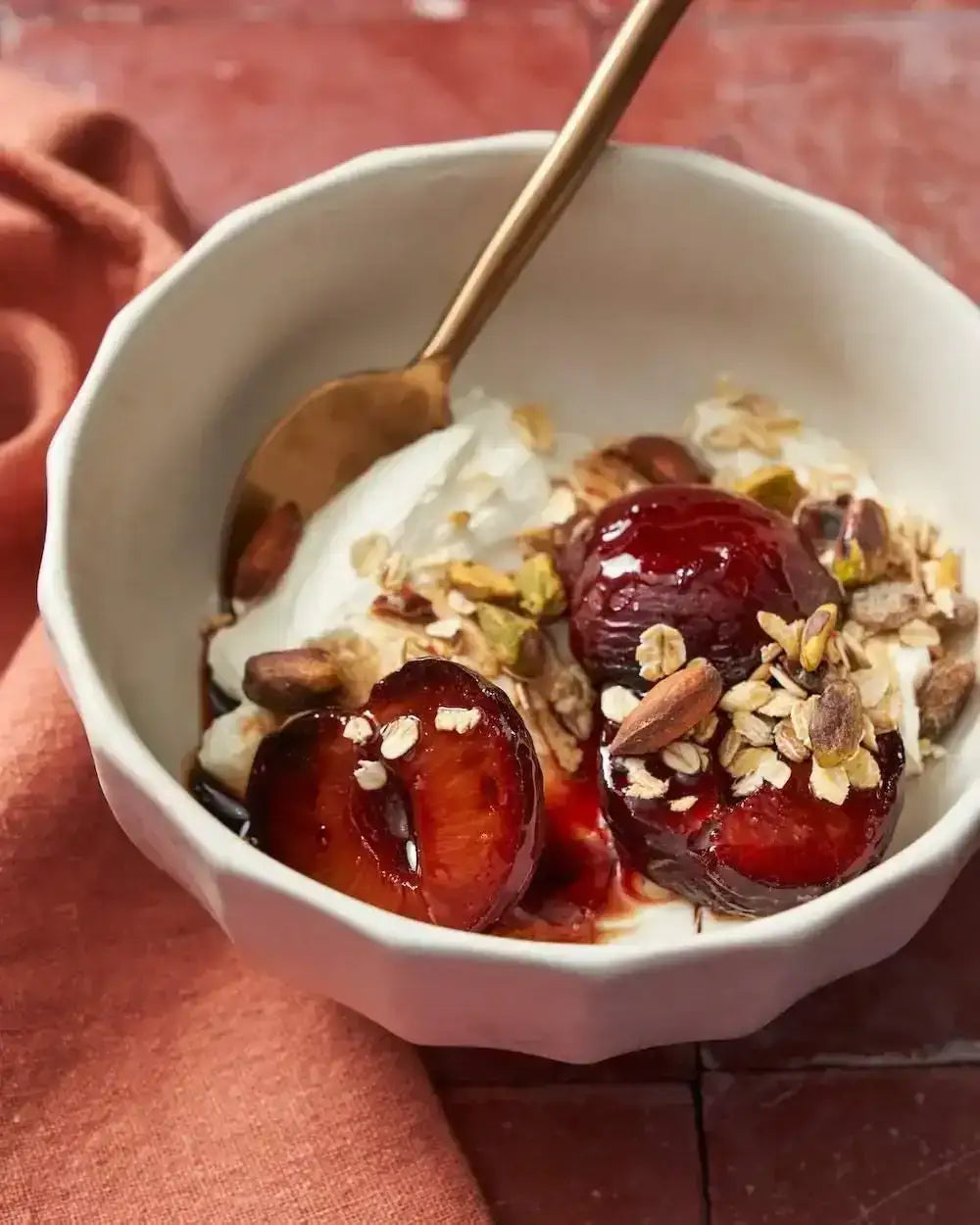 Asoke ceramic bowl with cream, roasted plums, oats, and nuts on terracotta surface with rust cloth