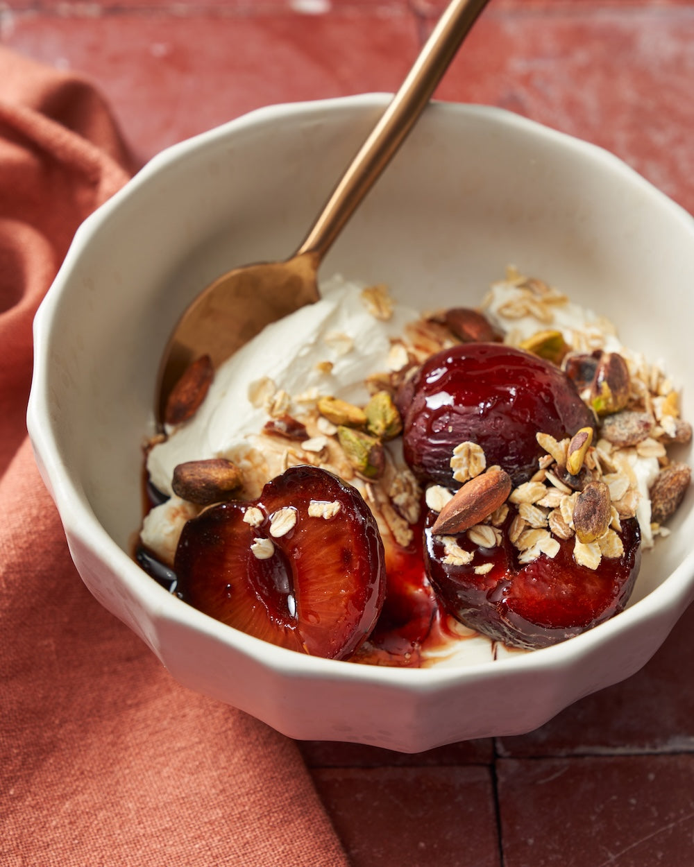 Ceramic bowl with roasted plums, granola, nuts, and yogurt on a rustic table.