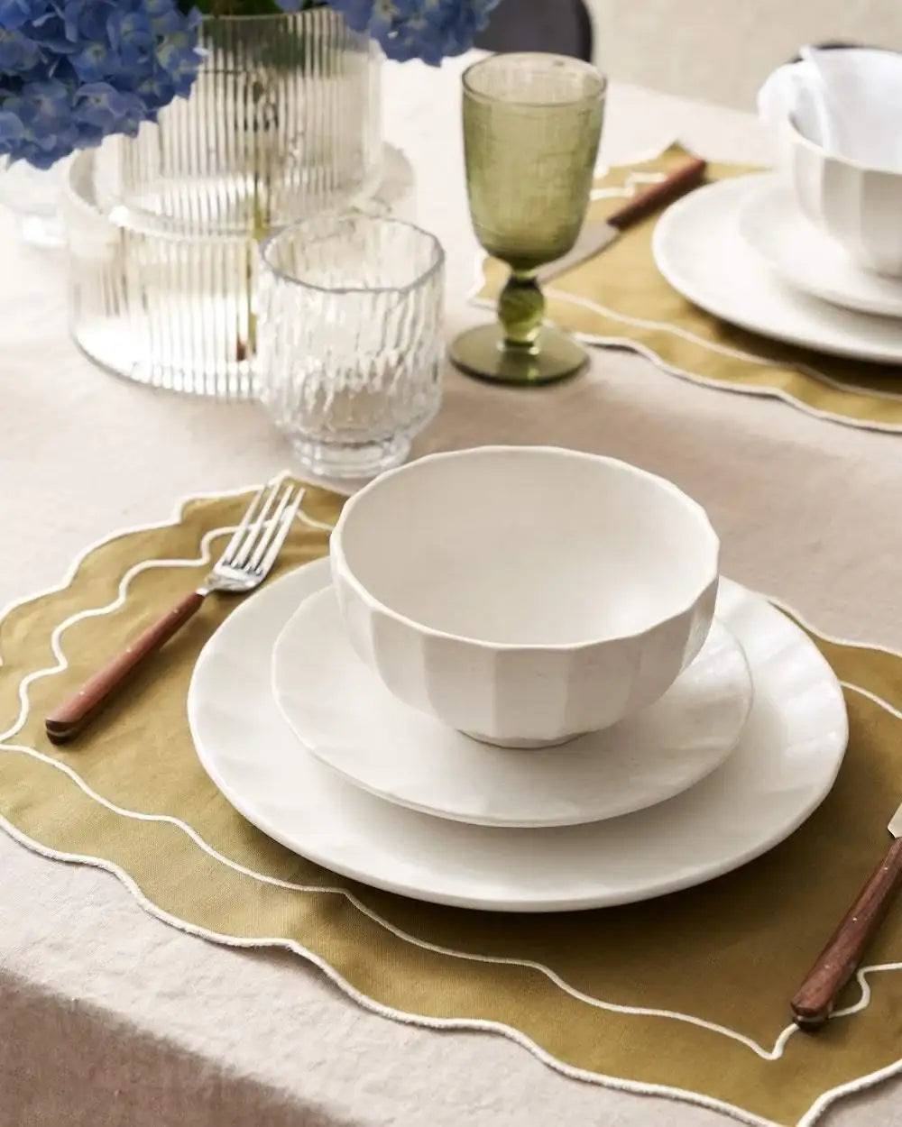 Minimalist white ceramic bowl and plates on olive green scalloped placemat with wooden handle cutlery, glassware, blue flowers