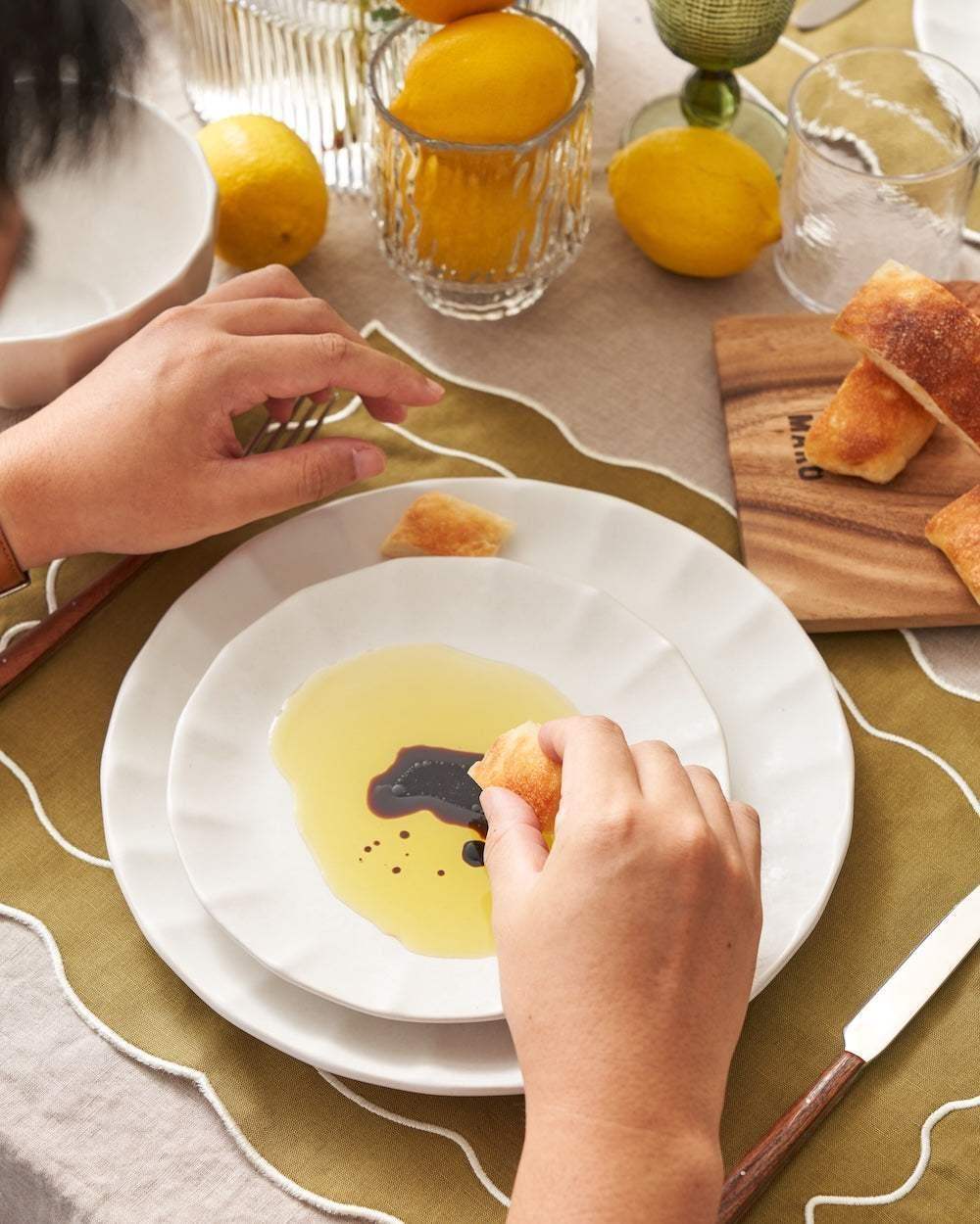 Hands dipping bread pieces in olive oil and balsamic vinegar on white Asoke ceramics plate by Marion Grasby