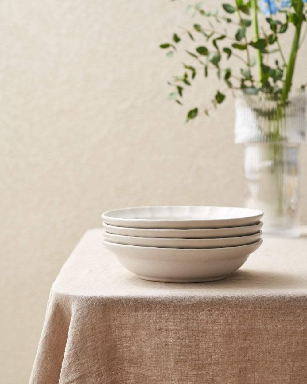 Stack of four white ceramic plates on beige linen tablecloth with green branches in glass vase