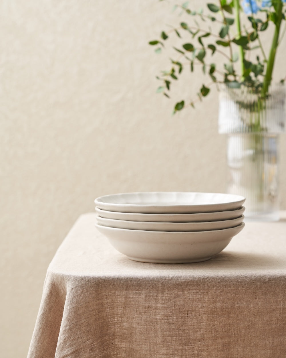 Stack of ceramic dinner bowls on a beige linen tablecloth with a glass vase and greenery