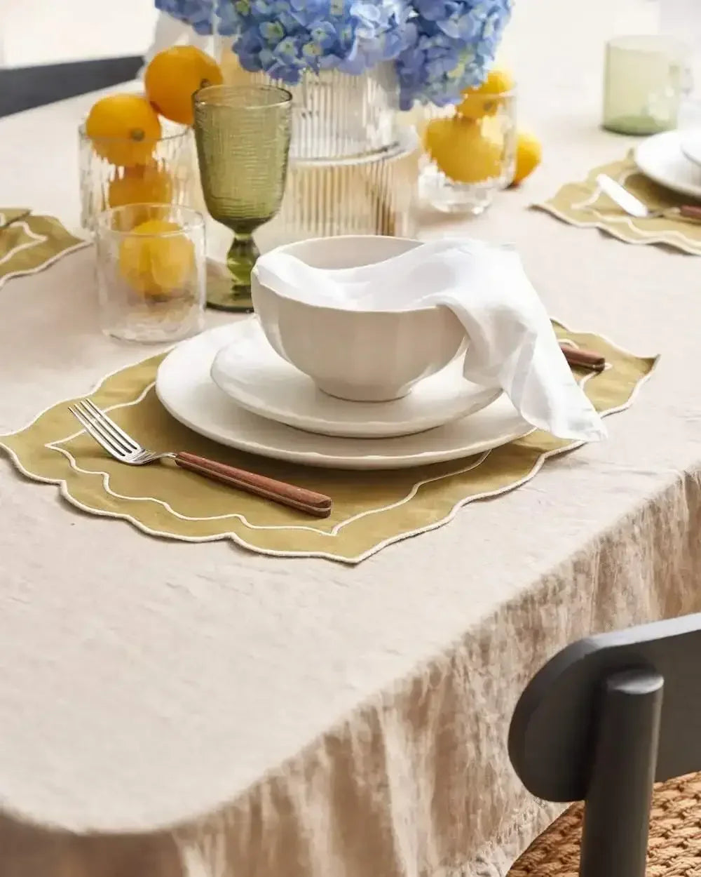 Elegant dining table setting with white ceramic bowl, plate, cloth napkin, yellow placemat, glassware, lemons, and blue hydrangeas.