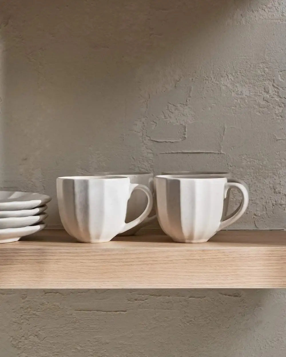 White ceramic cups and plates on a wooden kitchen shelf with textured beige wall backdrop