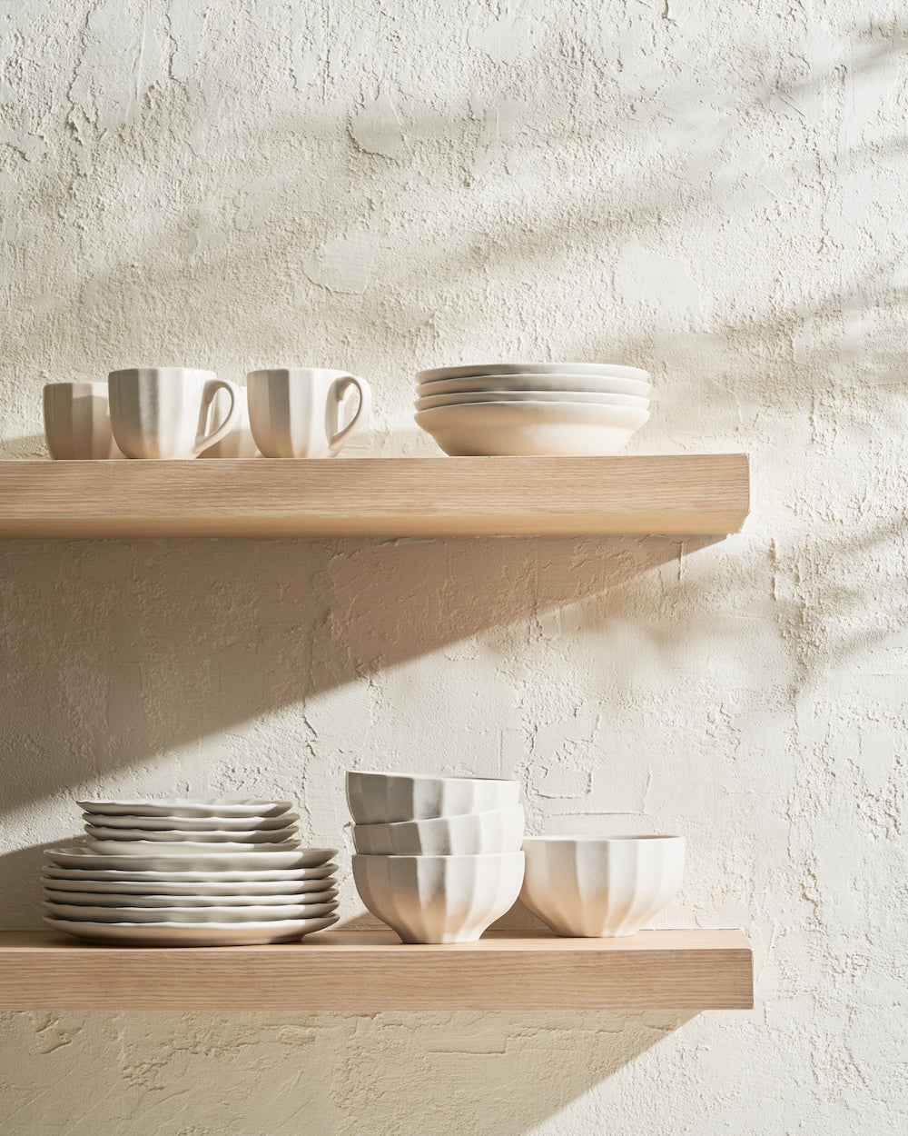 Minimalist ceramic mugs, bowls, and plates arranged on wooden kitchen shelves in natural light
