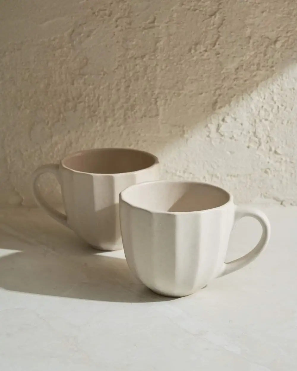 Two neutral-toned ceramic mugs with textured walls on a marble surface in natural kitchen light by Marion Grasby