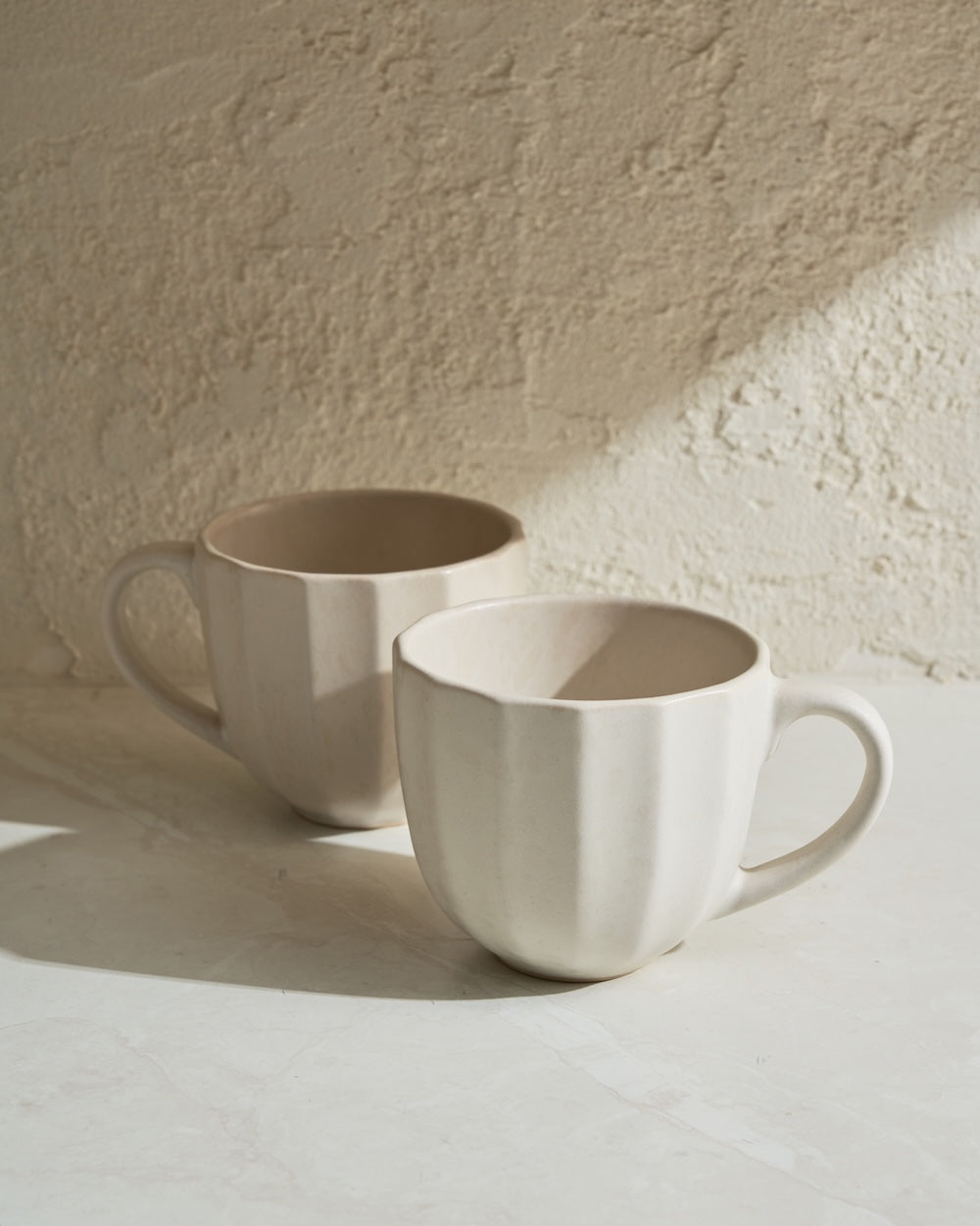 Two cream ceramic mugs with textured sides on a light stone surface in natural light.