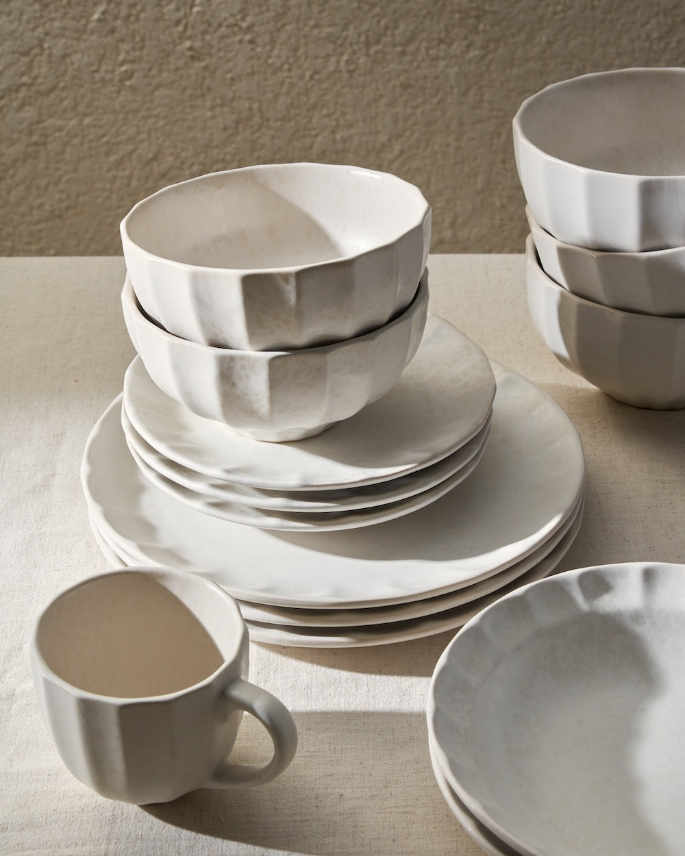 Stack of handmade white ceramic bowls, plates, and mug on neutral tabletop for tableware and dining.
