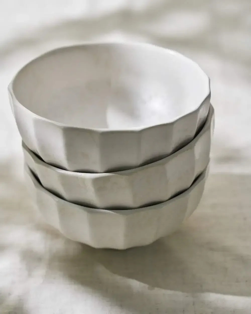 Stack of three white ceramic bowls with faceted texture on a neutral fabric surface
