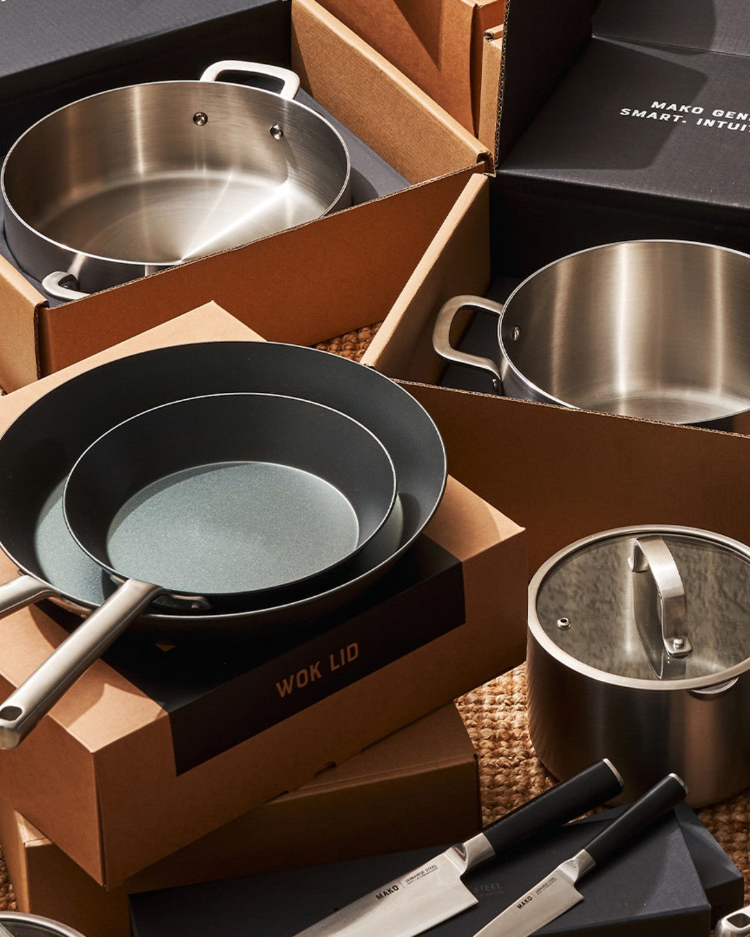 Set of MAKO cookware including stainless steel pots, black steel pans, and knives.