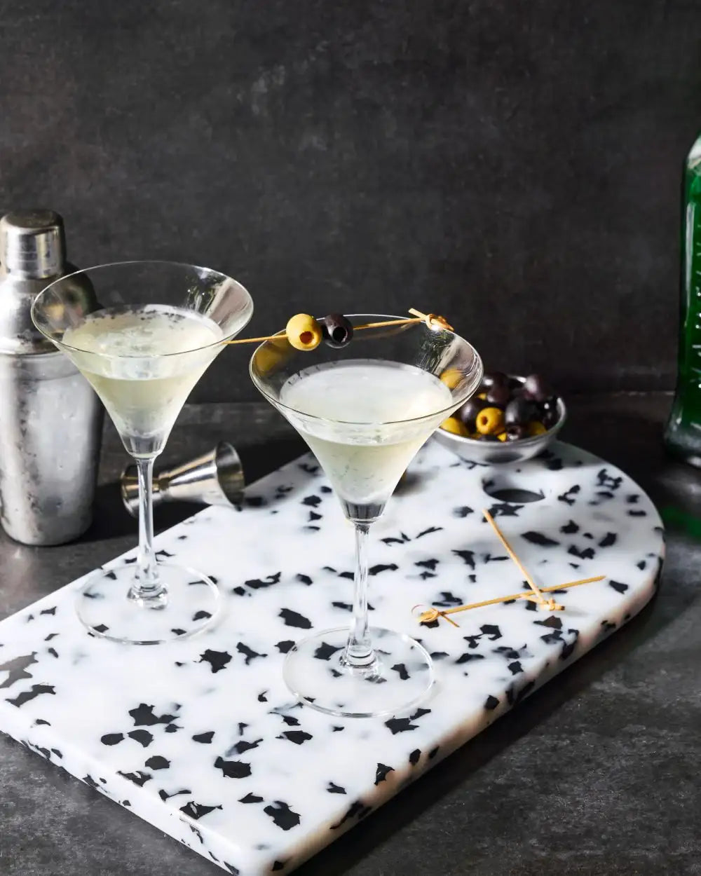 Two martini glasses with olives on a marble serving board, cocktail shaker and olives nearby