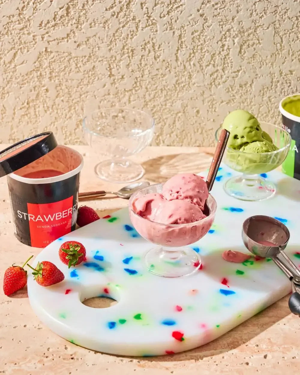 Strawberry and matcha ice cream in glass bowls with fresh strawberries on a colorful cutting board.