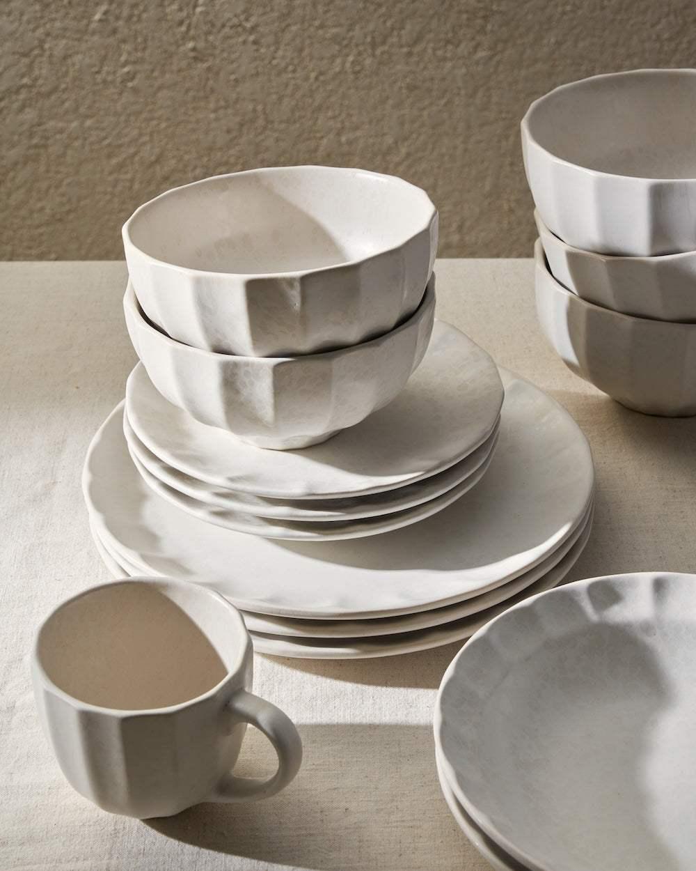 Modern white ceramic dinnerware set with bowls, plates, and mug on a neutral tablecloth.