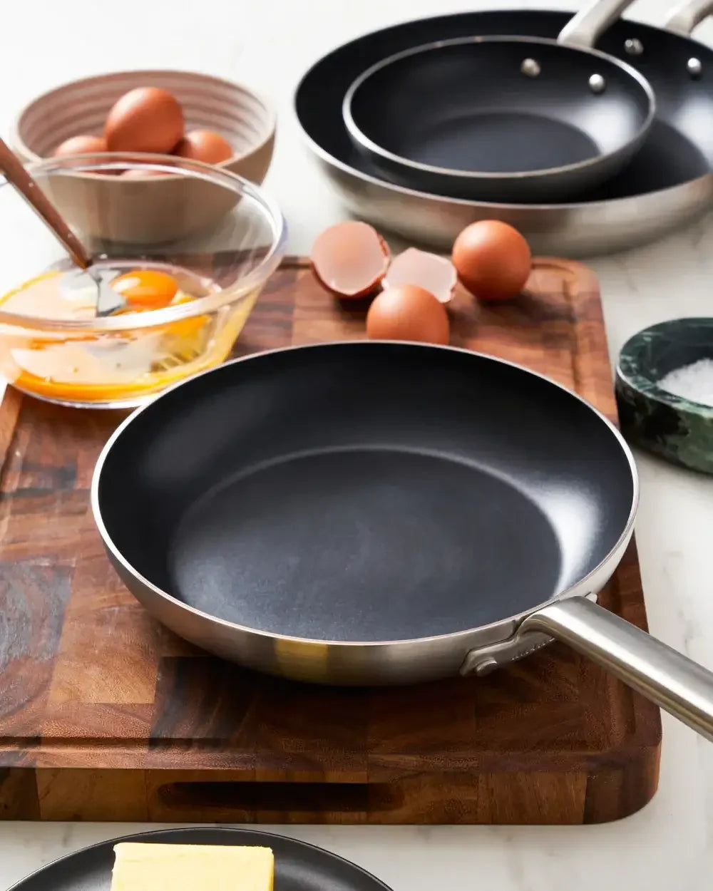 Non-stick frying pan on wooden board with eggs, butter, and mixing bowl in kitchen setting