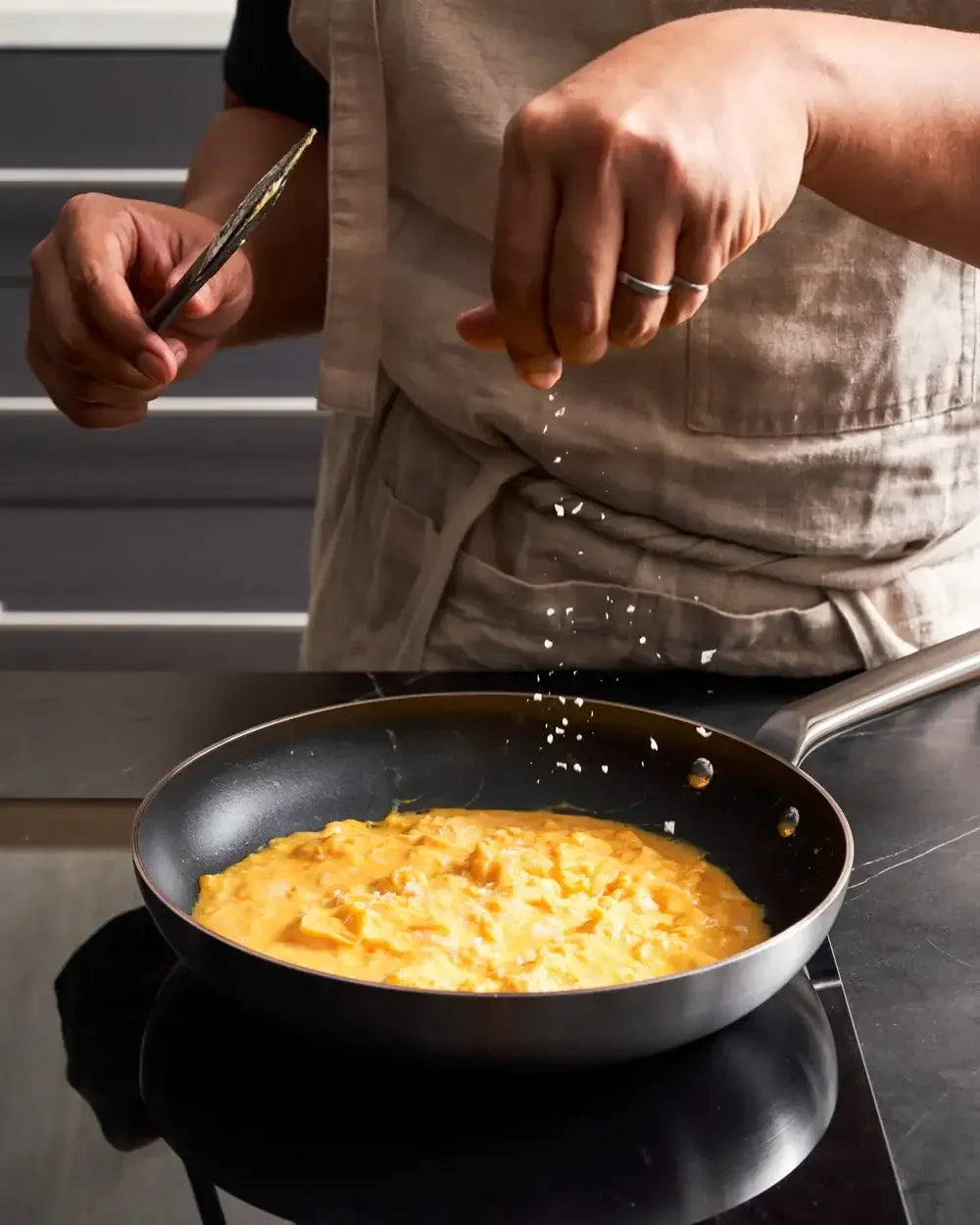 Hand seasoning scrambled eggs in a non-stick frying pan on a stovetop