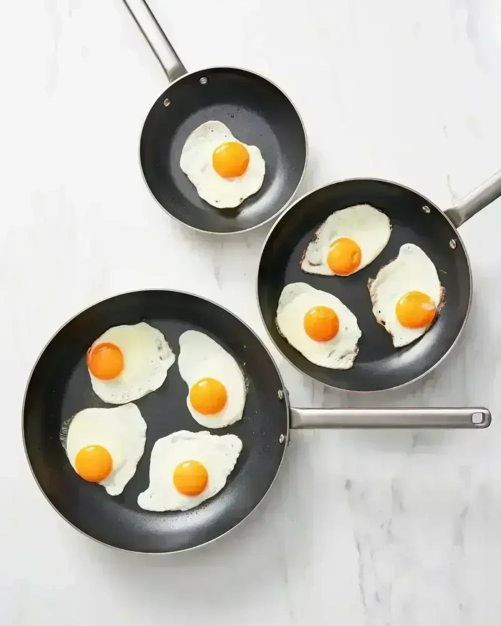Three non-stick frying pans with sunny side up eggs on a white countertop