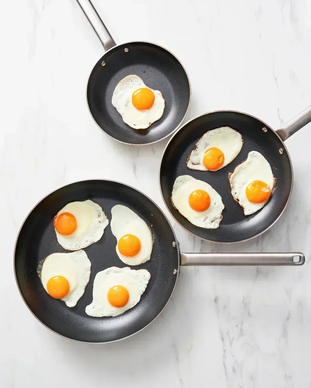 Three black non-stick frying pans with metal handles frying sunny-side-up eggs on a marble surface