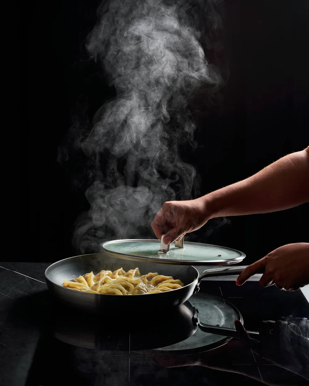 Hand lifting glass lid off non-stick frying pan with steamed dumplings over black stovetop
