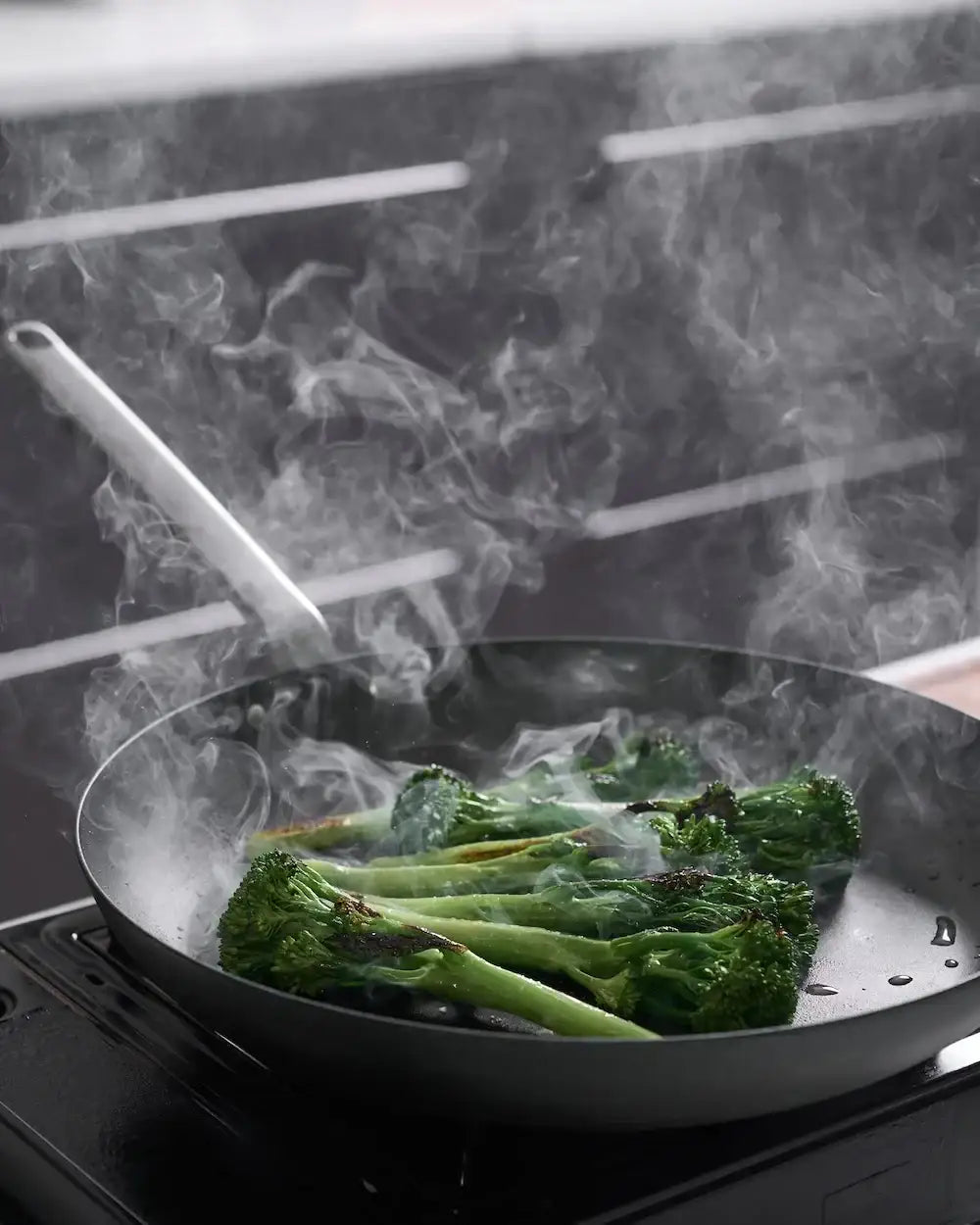 Black steel frying pan by Marion Grasby cooking charred broccolini on stove with rising steam