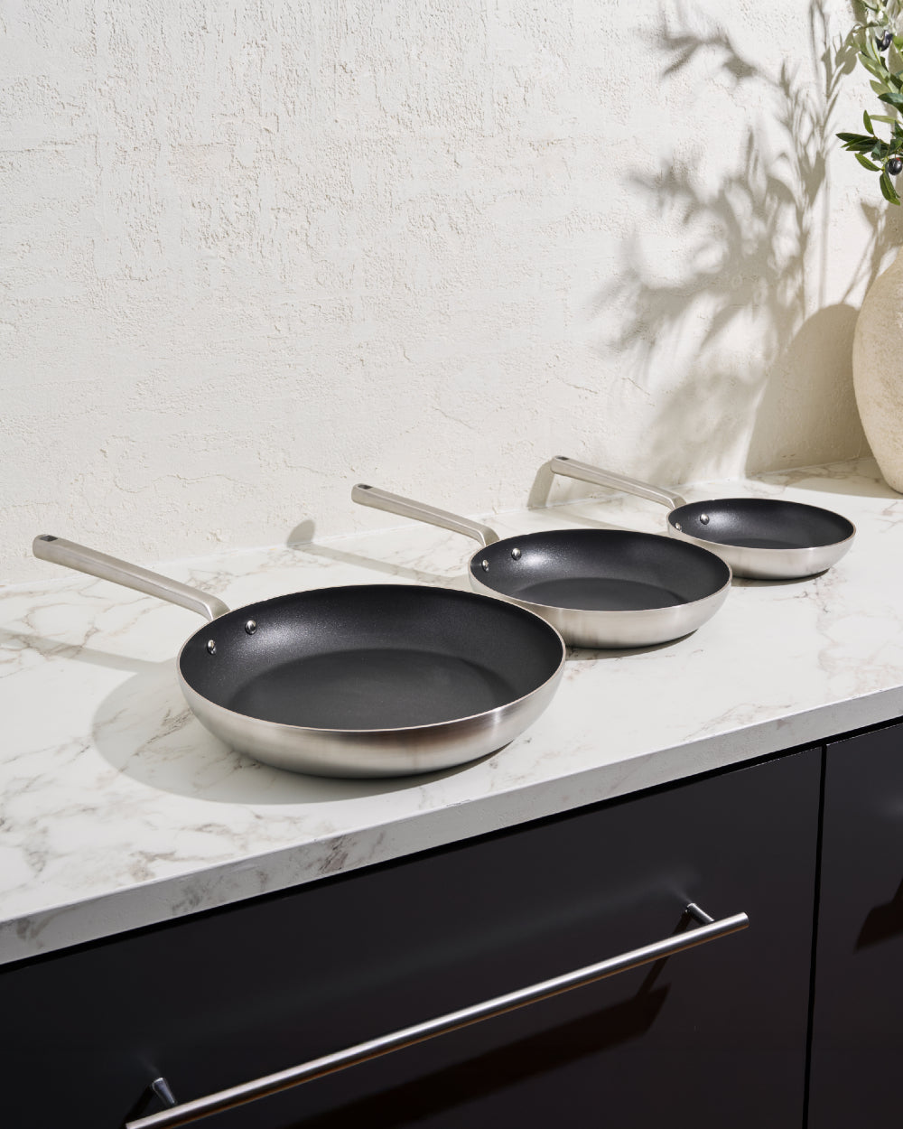 Set of three non-stick frying pans with stainless steel handles on marble kitchen counter