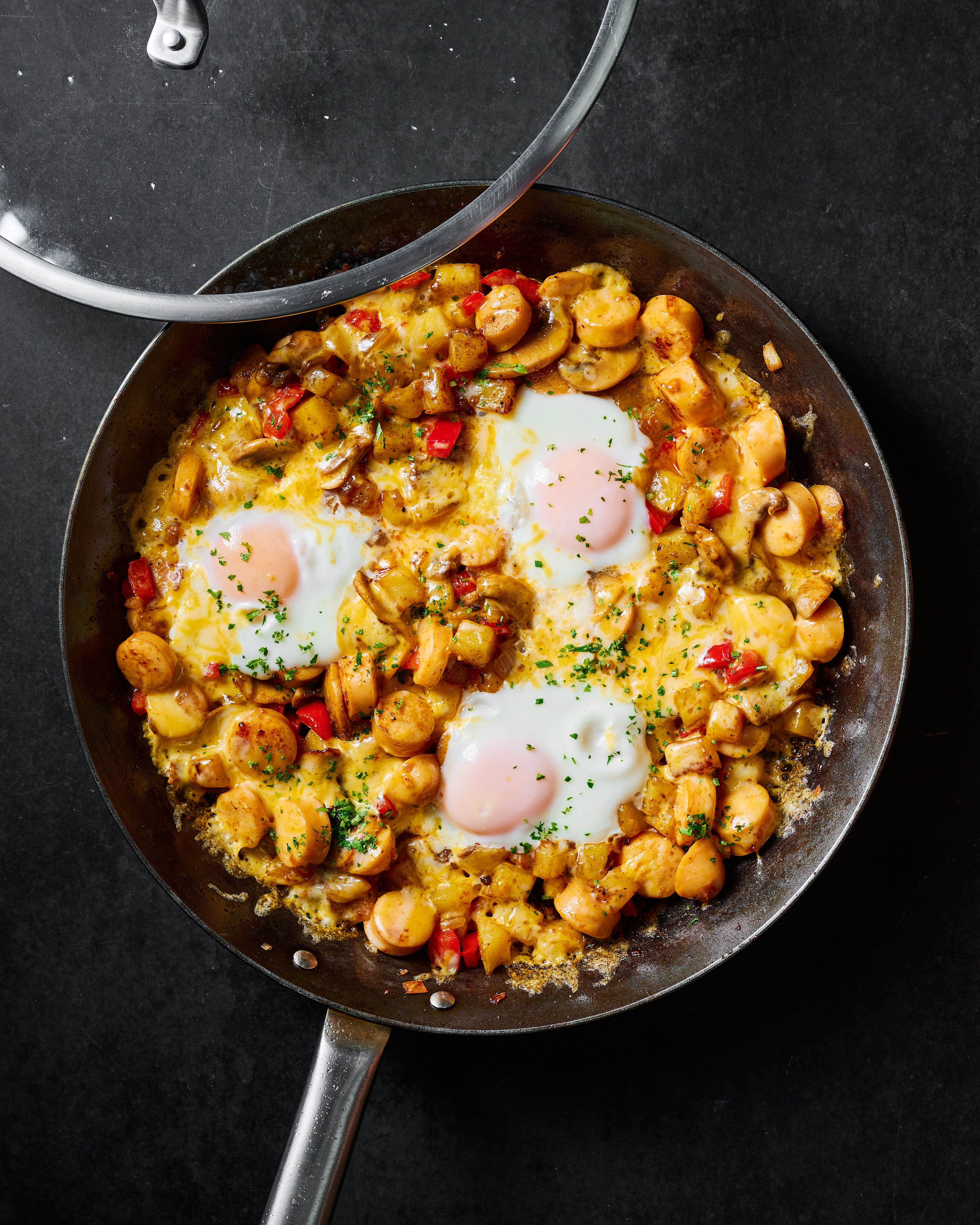 Overhead view of a skillet containing a creamy mushroom and chickpea breakfast dish with three sunny eggs on a dark background, with a glass lid partially visible.