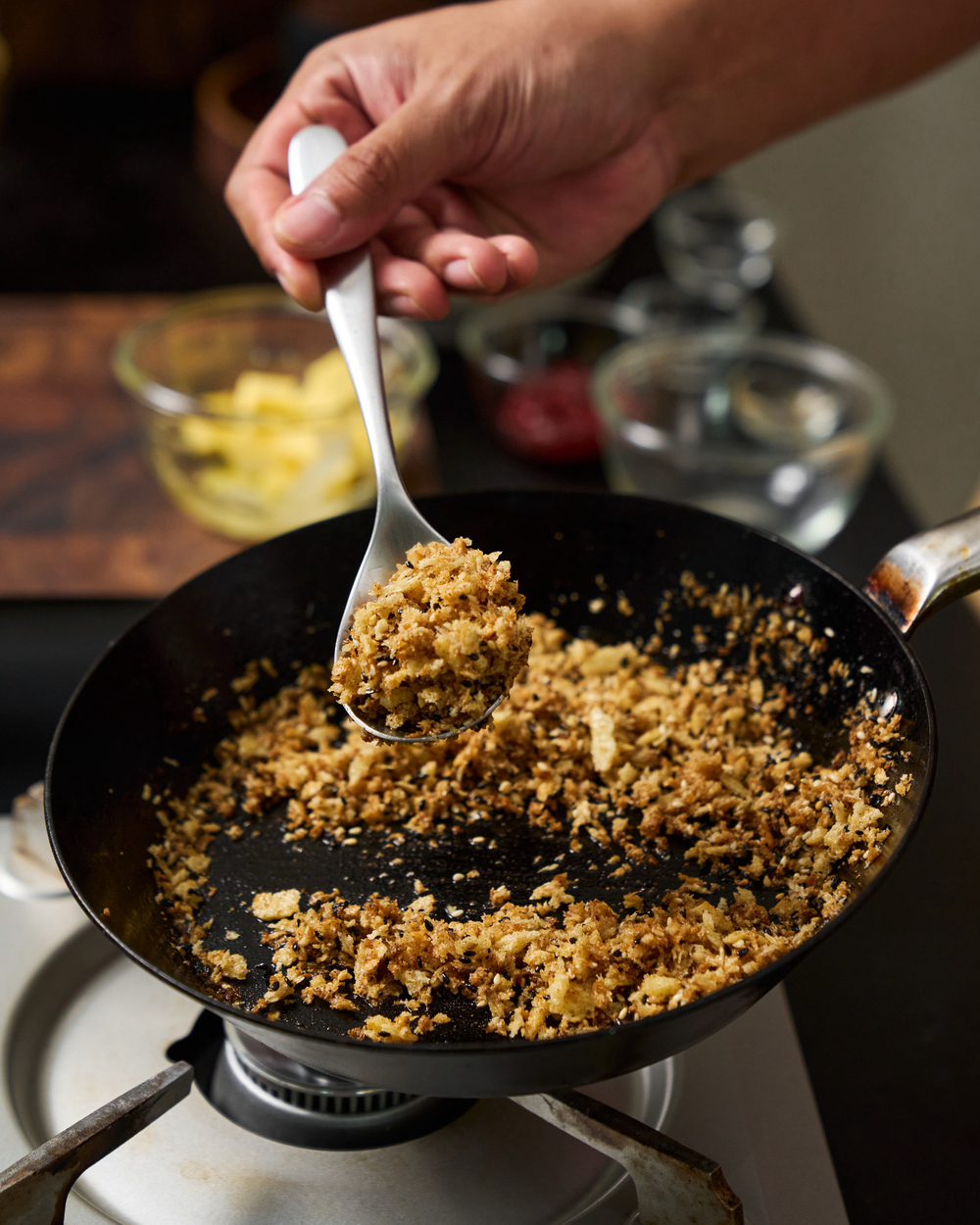 Golden panko breadcrumbs and sesame seeds being toasted in a black frying pan on a gas stovetop