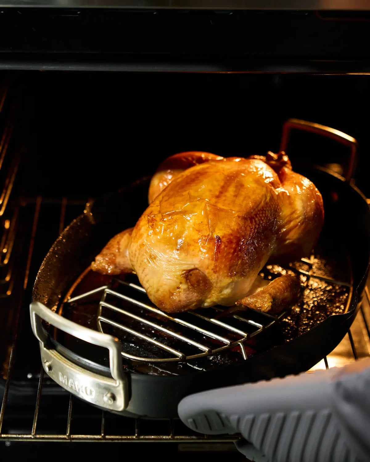 Whole chicken with golden, crispy skin roasting in a MAKO oval roasting pan inside the oven.