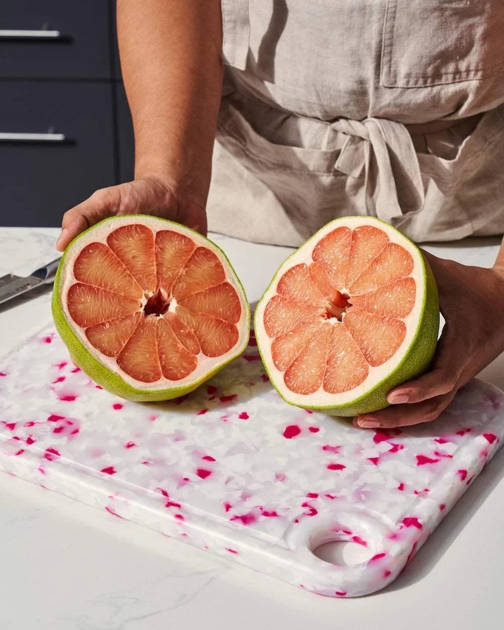 Person holding two halves of a fresh pink pomelo on a white and pink speckled cutting board in a kitchen