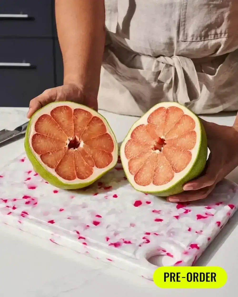 Person holding halved pomelo on white and pink speckled cutting board, pre-order sign