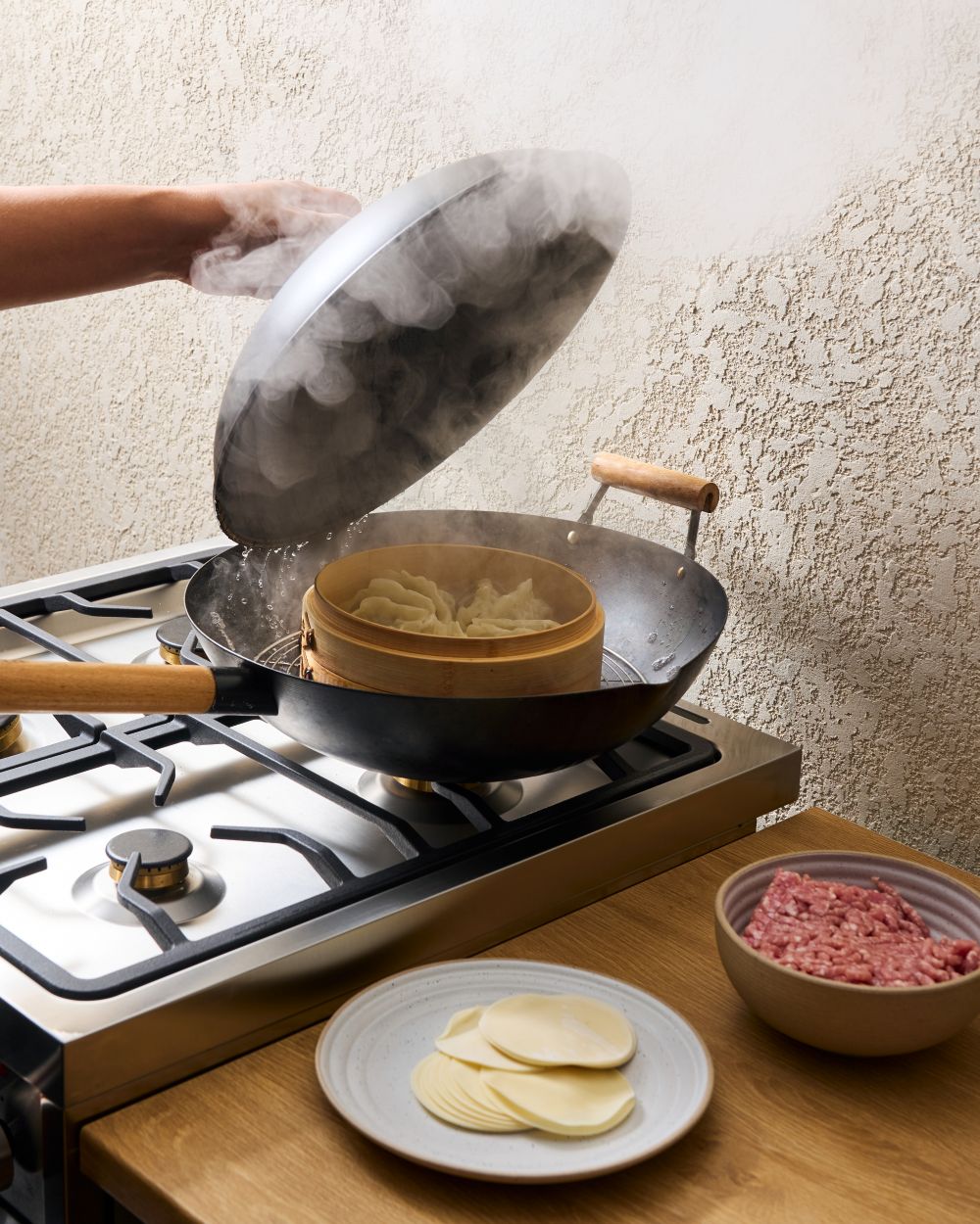 Steaming dumplings in a bamboo steamer on a gas stove with dough wrappers and ground meat nearby