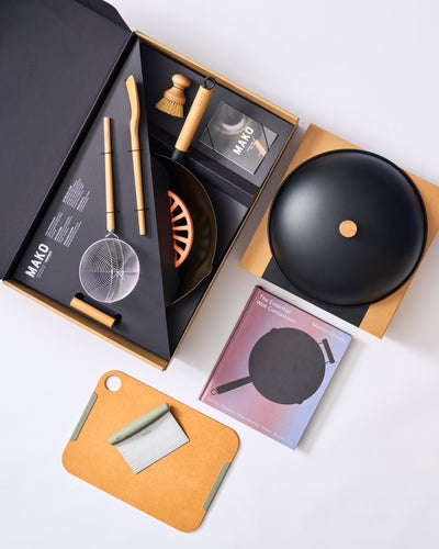Wok kit with black wok, utensils, cleaning brush, cookbook, and cutting board set on white background