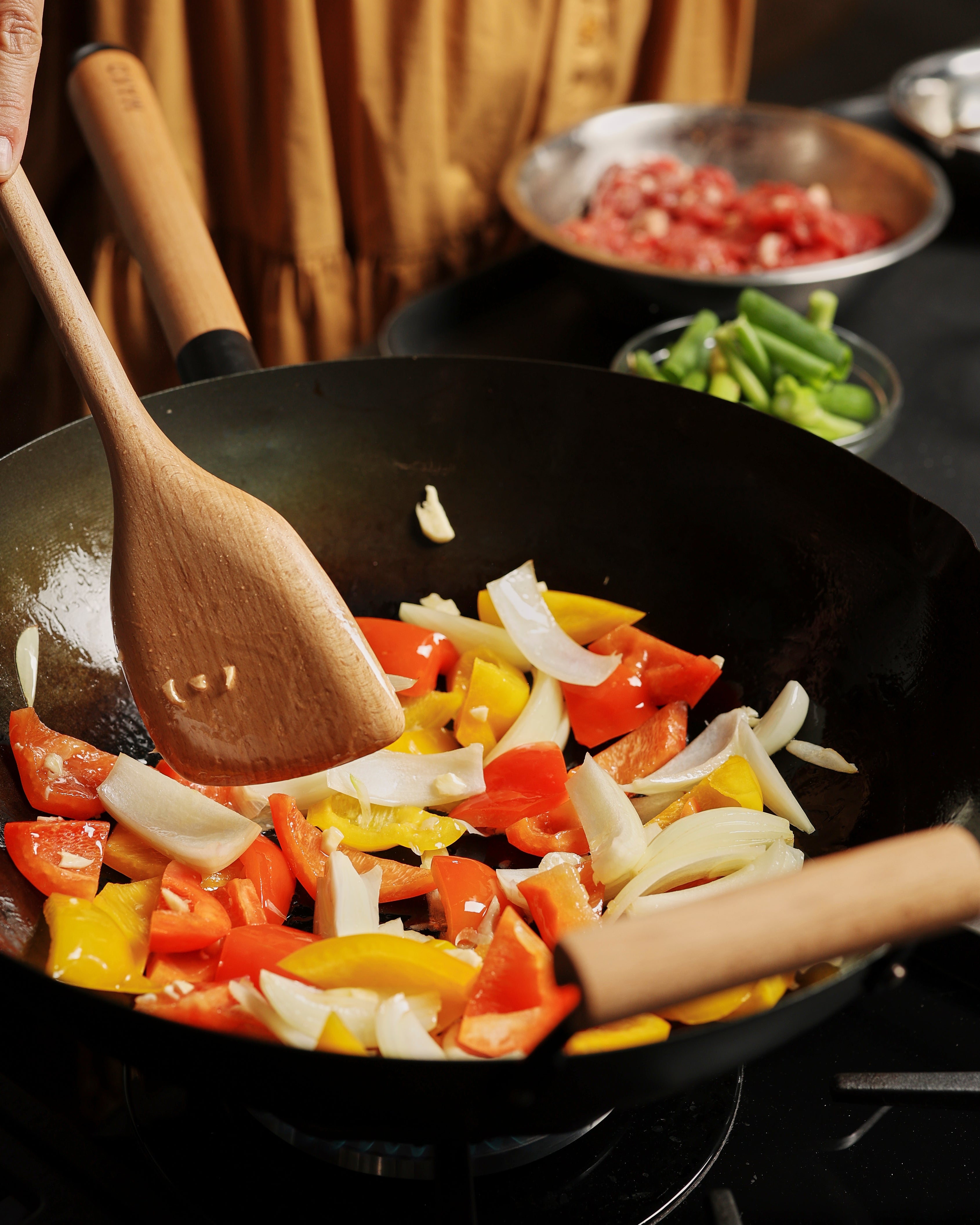 Stir-frying peppers and onions in a wok with wooden utensils, fresh ingredients in background