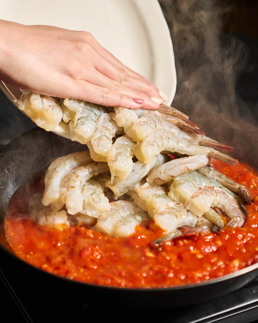Raw shrimp being added from a white plate into a black pan filled with red tomato sauce, with steam rising.