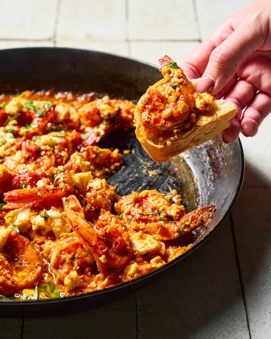 A close-up of a hand holding a slice of toasted bread topped with a generous serving of spicy shrimp and sausage bruschetta, with more of the same dish served in a large black pan on a tiled kitchen countertop.