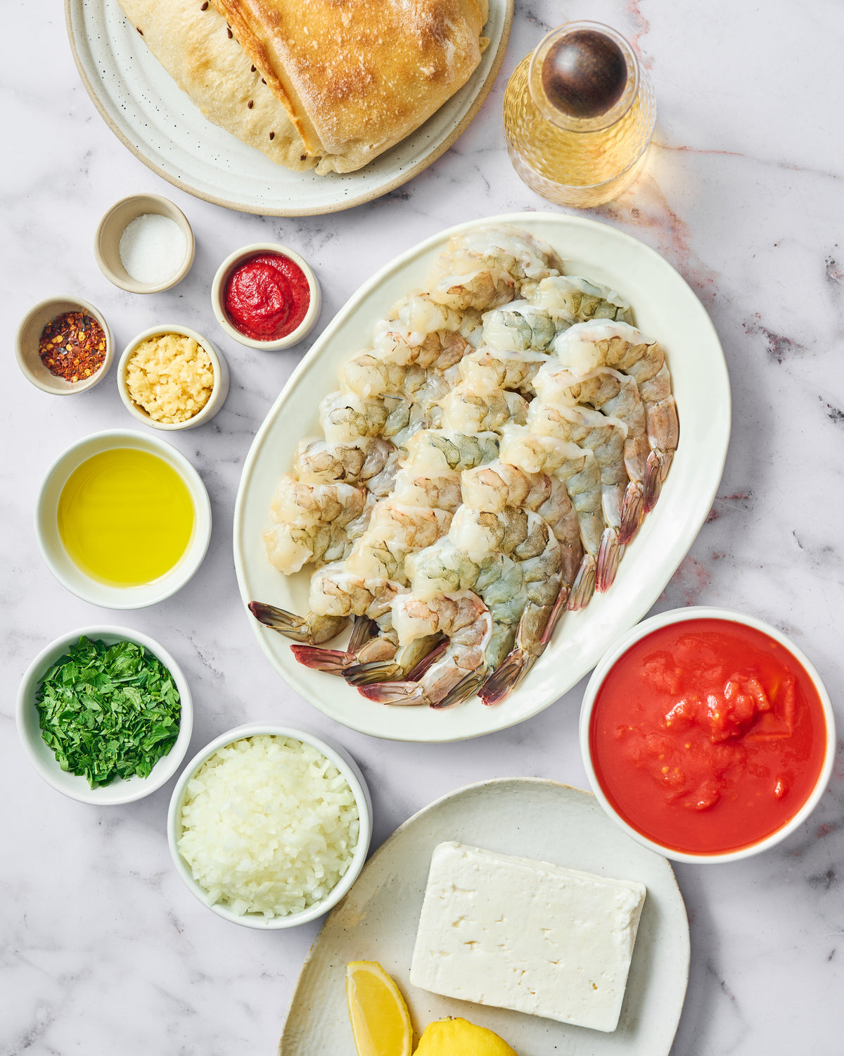 An overhead flat-lay of a seafood pasta recipe, featuring a large platter of raw shrimp, a block of tofu, chopped onions, parsley, garlic, red tomato sauce, olive oil, and spices, arranged on a white marble surface with a plate of pasta and bread nearby.