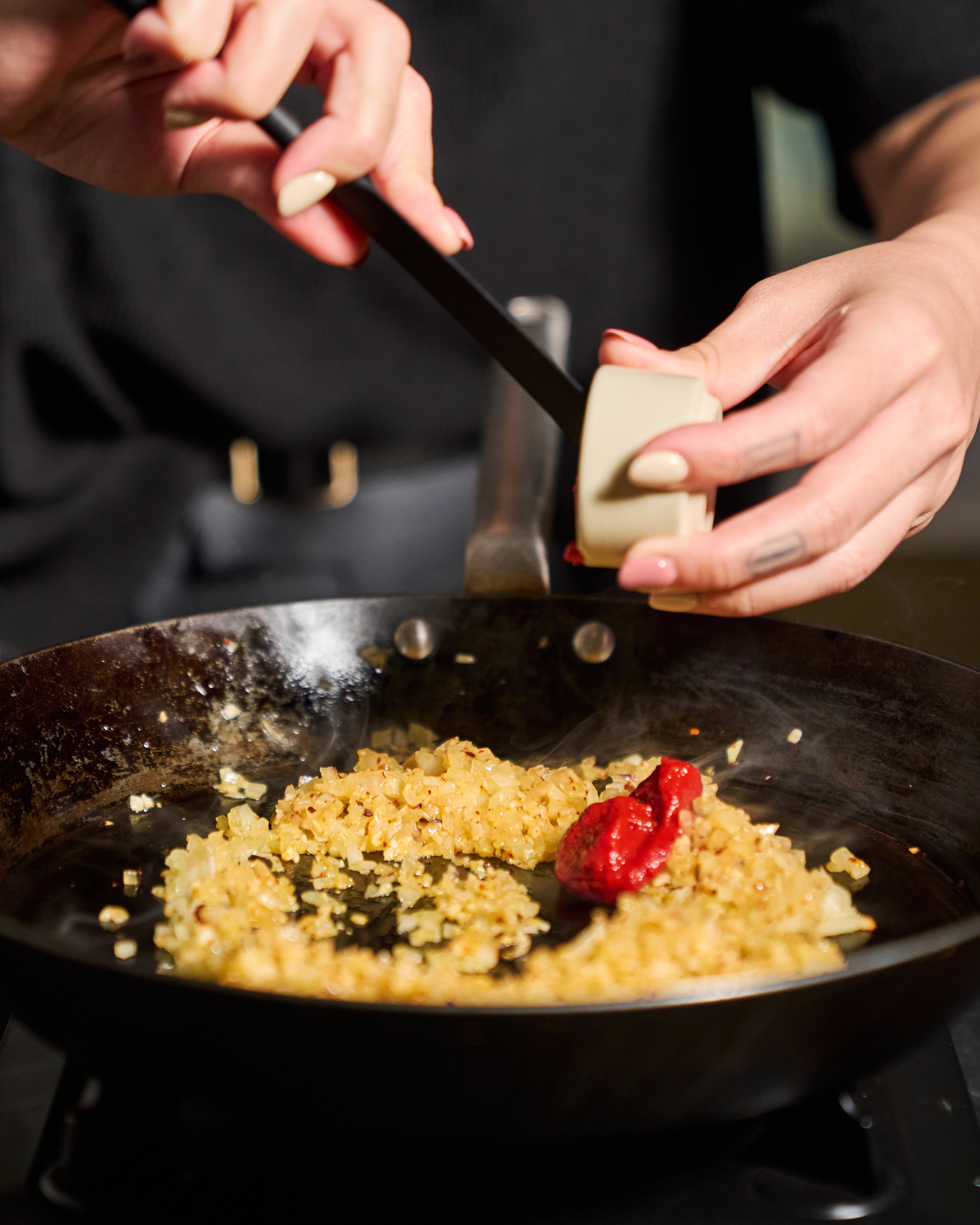 A close-up lifestyle shot of a person's hands adding a chunk of butter into a black frying pan with sautéed onions and a red pepper on a stove, set against a dark background.